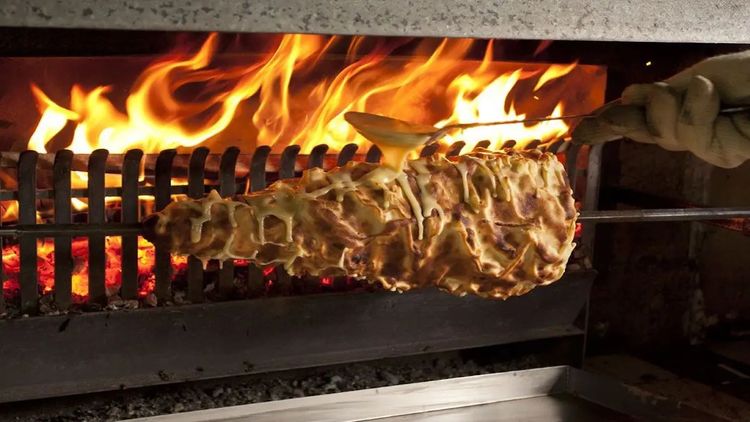 Une personne verse de la pâte pour réaliser un gâteau à la broche devant un feu de bois.