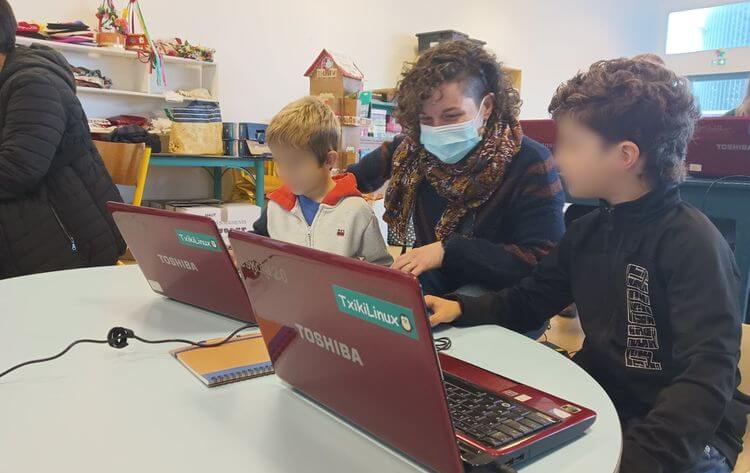Une adulte montre à deux enfants le logiciel Txikilinux sur des ordinateurs dans une salle de classe.
