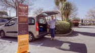 Photo du présentoir de l'entreprise et Vincent Chabbert tenant dans sa main la porte du coffre ouvert d'une voiture