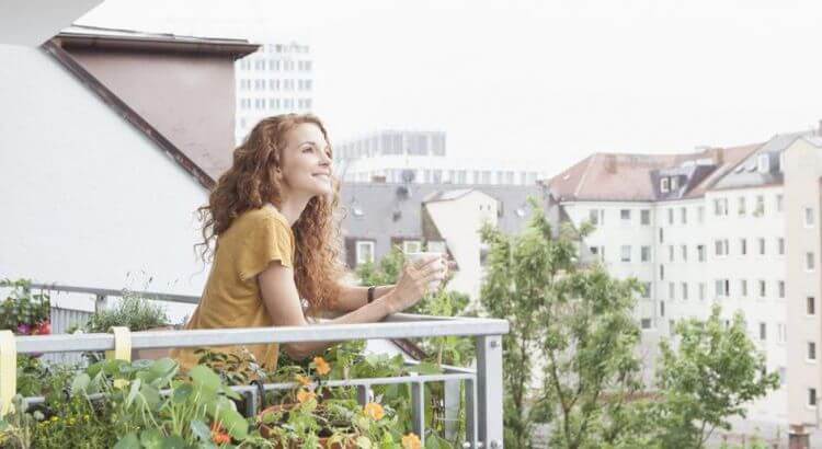 Une femme souriante tenant un verre d'eau sur un balcon accoudée à la rembarde