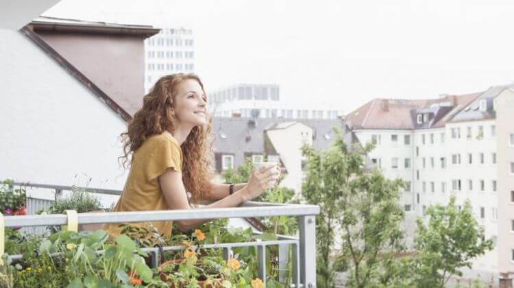 Une femme souriante tenant un verre d'eau sur un balcon accoudée à la rembarde
