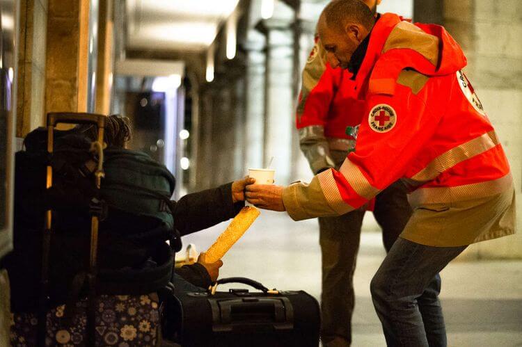 Bénévole de la Croix Rouge donnant une boisson chaude à une personne assise par terre