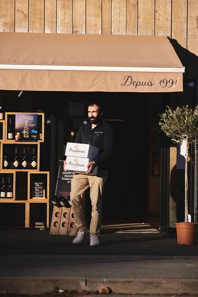 Julien Combes maître caviste au Pays basque, sortant de sa boutique avec 2 caisses de vin dans les bras
