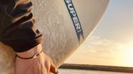 Iban Carrere porte l'un de ses bijoux au poignet, sur une plage, avec une planche de surf sous le bras.