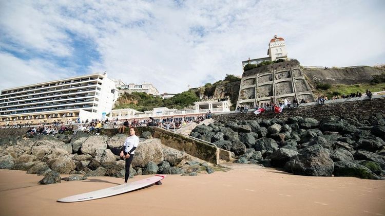 Un surfeur se prépare sur l'une des plages de Biarritz avec la ville et le public derrière lui.