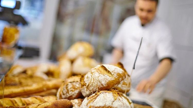 Des pains sont exposé en premier plan, devant un boulanger qui travaille.