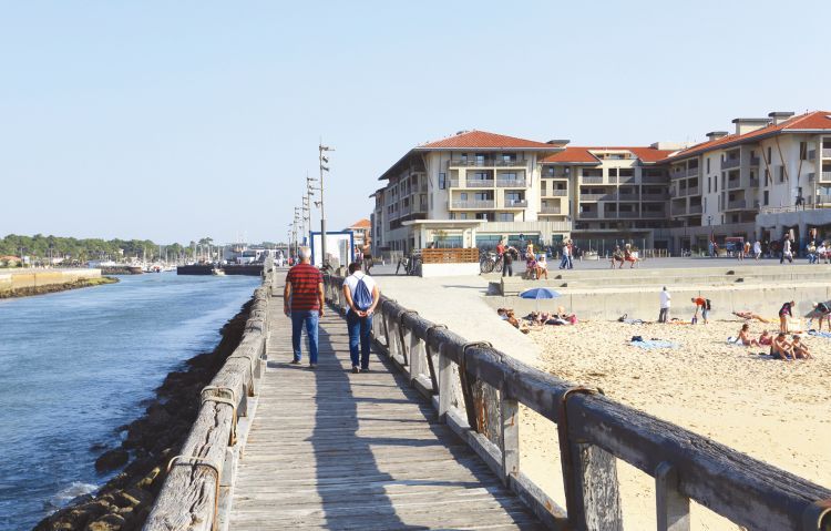 Une photo prise depuis une promenade sur le bord de l'eau, avec des résidences dans le fond, une plage au premier plan.