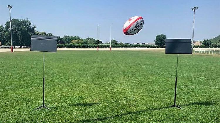 Un ballon de rugby qui passe au dessus d'un fil tendu entre deux pupitres de musique posés sur un terrain