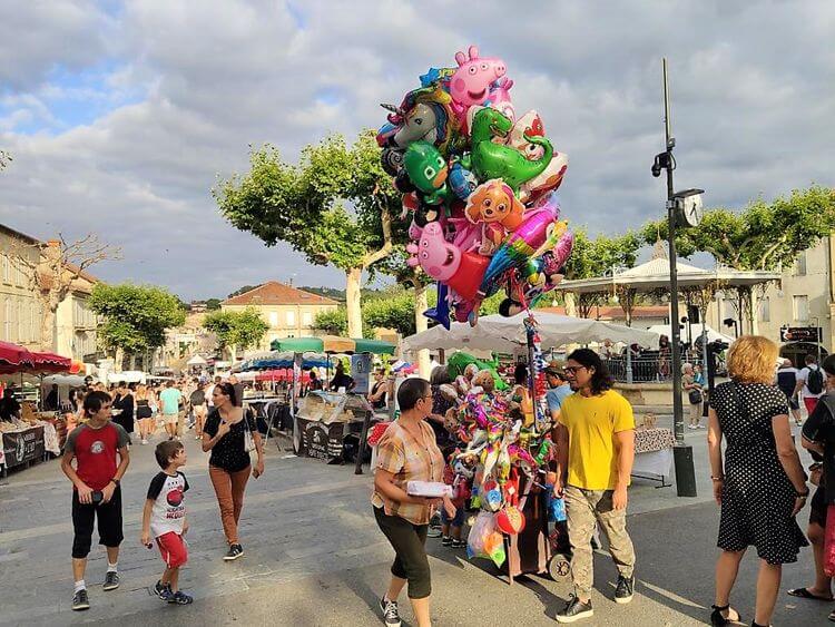 Un marchand de ballons sur la place du kiosque et des gens qui se promènent