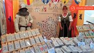 Anita Montagne et un homme en armure derrière le stand de l'association Histoire de Transmettre avec des blasons, une carte de France, des objets à la vente