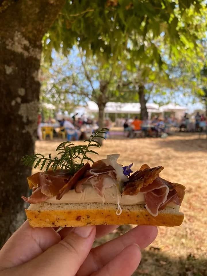 Une main qui tient une tartine avec du jambon et une préparation à l'ail avec des personnes en fond en train de pique-niquer sous des arbres